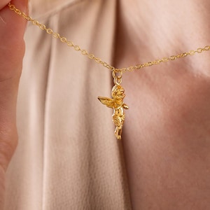 May include: A gold-colored necklace featuring a small cherub pendant. The cherub has outstretched wings and is suspended from a delicate chain. The necklace is being held up against a neutral background.