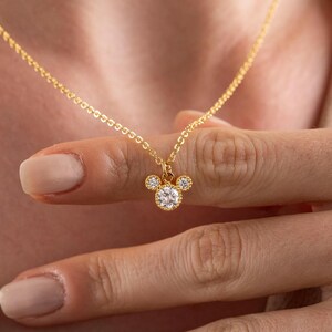 Peut inclure: Un collier doré avec un pendentif en forme de tête de souris. Le pendentif est orné d'une grosse pierre précieuse ronde et transparente au centre, avec deux petites pierres précieuses au-dessus. Le collier est tenu par une personne.