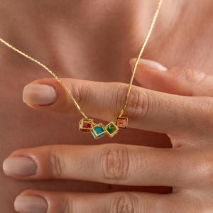 May include: A gold-colored necklace featuring a series of small, colorful square pendants. The pendants are in shades of red, turquoise, green, and orange. The necklace is delicate and appears to be worn around the neck.