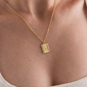 Könnte beinhalten: Eine goldfarbene Halskette mit einem rechteckigen Anhänger mit dem Buchstaben "A". Der Anhänger ist von kleinen, dekorativen Details umgeben. Die Halskette wird über einem weißen Oberteil getragen.