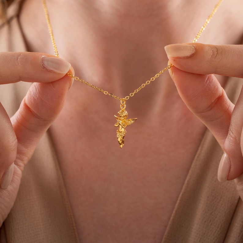 May include: A gold-colored necklace featuring a small cherub pendant. The cherub has outstretched wings and is suspended from a delicate chain. The necklace is held up against a neutral background, highlighting the jewelry's design and color.