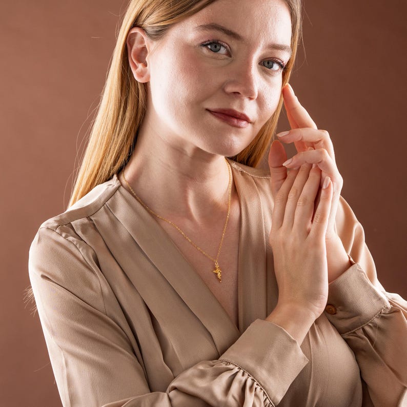 May include: A gold-colored necklace with a small pendant is worn by a person. The pendant is a detailed, possibly floral, design. The person is wearing a beige satin blouse. The background is a solid brown color.