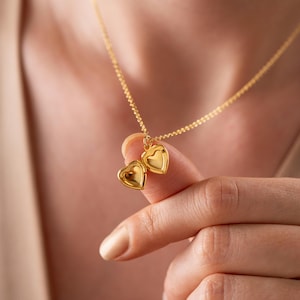 Pode incluir: Um colar dourado com dois pingentes em forma de coração. Os pingentes estão suspensos por uma corrente fina. O colar é segurado pelos dedos de uma pessoa, destacando o design e a cor da joia.