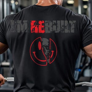 Puede incluir: Una camiseta negra de manga corta con el texto I'M REBUILT en letras grises y rojas desgastadas en la parte superior de la espalda. Debajo del texto hay un gr&aacute;fico circular rojo de cara sonriente con un dise&ntilde;o de calavera cosida.