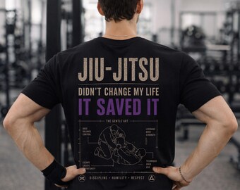 May include: A black t-shirt featuring a graphic on the back with the text JIU-JITSU DIDN'T CHANGE MY LIFE IT SAVED IT in beige and purple. Below the text is a line drawing of two people grappling, surrounded by labels and the words THE GENTLE ART.