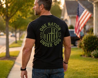 May include: A black short-sleeved t-shirt featuring a circular olive green graphic on the back. The design includes the text ONE NATION UNDER GOD, ESTD. 1776, and a stylized American flag with a cross integrated into the vertical stripes.