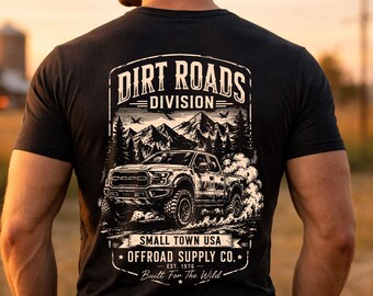 May include: Dark gray t-shirt featuring a graphic of a truck driving on a dirt road, with the text "DIRT ROADS DIVISION" and "OFFROAD SUPPLY CO." The design includes mountains, trees, and the phrase "Built For The Wild."