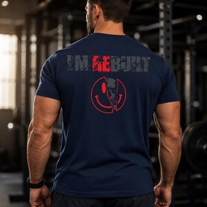 Puede incluir: Una camiseta azul marino de manga corta con el texto "I'M REBUILT" en una fuente desgastada en la parte superior de la espalda. Debajo del texto hay un gr&aacute;fico rojo de una cara sonriente, con la mitad derecha mostrando un dise&ntilde;o de calavera mec&aacute;nica.
