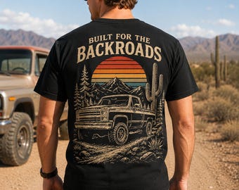 May include: Black t-shirt with a graphic of a truck, mountains, and a sunset. The text "BUILT FOR THE BACKROADS" is arched above the graphic. The shirt is worn by a person in a desert landscape.