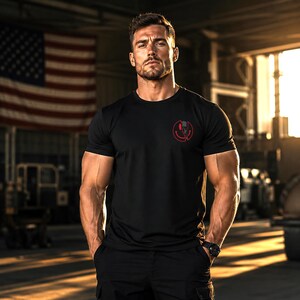 Puede incluir: Una camiseta negra de manga corta con un peque&ntilde;o gr&aacute;fico circular rojo en el pecho izquierdo. El gr&aacute;fico muestra un dise&ntilde;o dividido con una cara sonriente en un lado y una calavera en el otro. La camiseta tiene cuello redondo y corte ajustado.