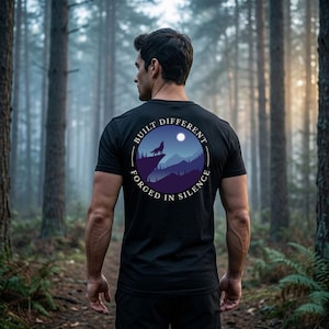 May include: Black t-shirt with a circular graphic featuring a howling wolf on a mountain, a full moon, and the text "BUILT DIFFERENT FORGED IN SILENCE". The shirt is worn by a person in a forest setting.