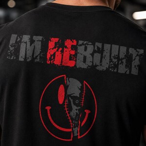 Puede incluir: Una camiseta negra con el texto "I'M REBUILT" en una fuente desgastada, con la palabra "RE" en rojo y el resto en gris. Debajo del texto hay un gr&aacute;fico de cara sonriente circular roja, cuya mitad es un dise&ntilde;o de calavera mec&aacute;nica cosida.