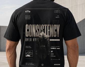 May include: Black t-shirt with the word "CONSISTENCY" in large, bold, white letters. Below it, the phrase "OVER HYPE" is printed. The shirt also features graphic elements and text including "DISCIPLINE, REPETITION, RESILIENCE".