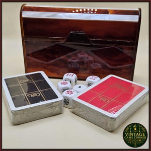 May include: A vintage game set featuring a brown, patterned box with a white accent. The set includes two decks of cards, dice, and a decorative logo. The cards are red and black. The dice are white with red markings.