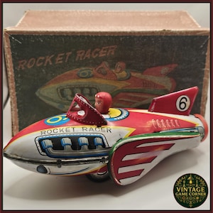 May include: Vintage tin toy rocket racer in red, white, and silver with a driver figure. The toy has the words "ROCKET RACER" printed on the side and a number 6 on the tail fin. The original box is in the background.