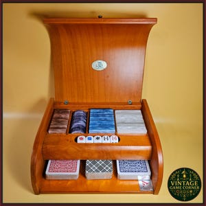 Op de afbeelding: Een houten spelopbergdoos met een open deksel voorzien van een metalen embleem. Het interieur bevat vier vakken gevuld met stapels gekleurde fiches, een rij van vijf witte dobbelstenen en drie kaartspellen met patronen op de achterkant.