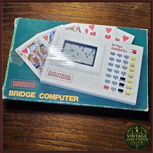 May include: A vintage Diamond Bridge Computer game. The white console has a screen and buttons, and is on a teal box. Playing cards with hearts and a King of Hearts are visible.