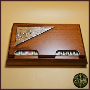 Könnte beinhalten: Eine rechteckige Spieleschachtel aus Holz mit einer polierten, silberfarbenen dreieckigen Einlage in der oberen linken Ecke, die mit einem Joker-Spielkartenmotiv geprägt ist. Die Vorderkante zeigt eine Reihe cremefarbener Würfel mit schwarzen und farbigen Augen.