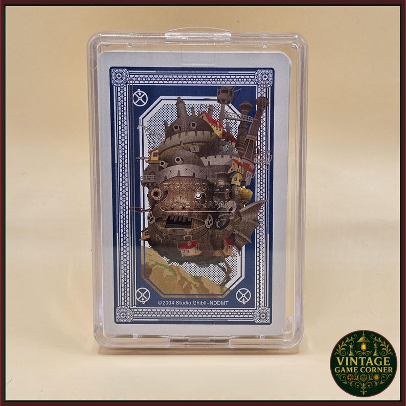 May include: A playing card featuring a detailed illustration of a moving castle from the animated film. The card is encased in a clear plastic case. The card has a blue border with white accents and the text "&copy;2004 Studio Ghibli - ND DMT".