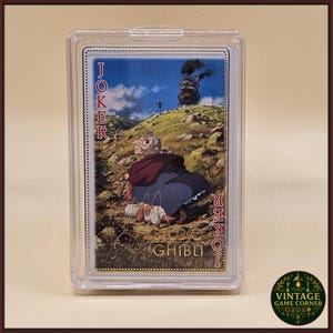 May include: A Studio Ghibli Joker playing card featuring an illustration of a character from the movie Howl's Moving Castle. The card is in a protective case. The word "Joker" is printed vertically on the left and right sides of the card.