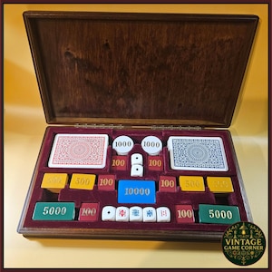 May include: A wooden game box with a hinged lid containing two decks of playing cards with red and blue backs, white circular chips marked 1000, red square chips marked 100, yellow chips marked 500, a blue chip marked 10000, and green chips marked 5000.