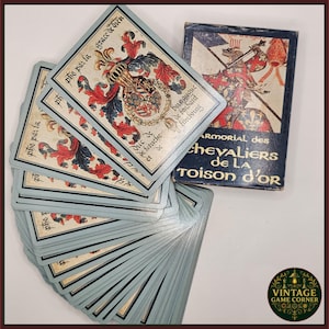 Könnte beinhalten: Ein Kartenspiel im Vintage-Stil mit hellblauem Rand und detaillierten Illustrationen. Die Karten zeigen Text und heraldische Designs in Rot, Blau und Gold. Die Kartenbox ist dunkelblau mit einer Ritterillustration und dem Text "ARMORIAL des CHEVALIERS DE LA TOISON D'OR."
