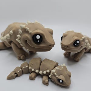 May include: Three tan-colored, articulated lizard figurines with white accents. The lizards have large, black and white eyes and a smiling expression. One lizard is segmented, and the other two are whole.