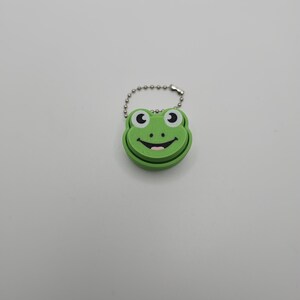 May include: A green frog-shaped keychain with a smiling face, black eyes, and a pink mouth. The keychain is attached to a silver ball chain. The frog's face is a bright green color.