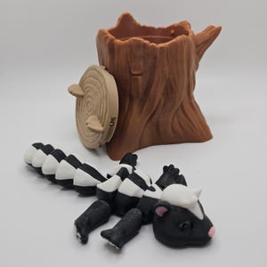 May include: A brown tree stump-shaped container with a hinged, circular lid. A black and white articulated skunk toy is in front of the container. The skunk has a white head with pink ears and a black stripe down its back.