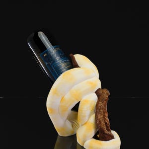 May include: A white and yellow snake-shaped wine bottle holder. The snake is coiled around a brown wooden support, holding a dark wine bottle. The background is black, highlighting the unique design.