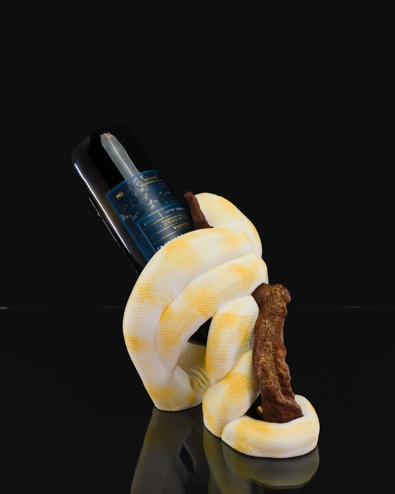 May include: A white and yellow snake-shaped wine bottle holder. The snake is coiled around a brown wooden support, holding a dark wine bottle. The background is black, highlighting the unique design.