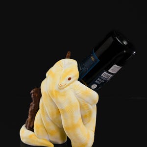 May include: A yellow and white snake-shaped wine bottle holder. The snake is coiled around a dark brown branch, supporting a black wine bottle. The snake has red eyes and a textured pattern.