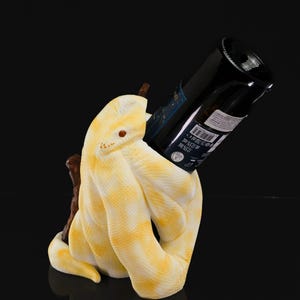 May include: A yellow and white snake-shaped wine bottle holder. The holder is designed to cradle a wine bottle, with the bottle angled upwards. The snake has a detailed scale pattern and a brown accent piece.