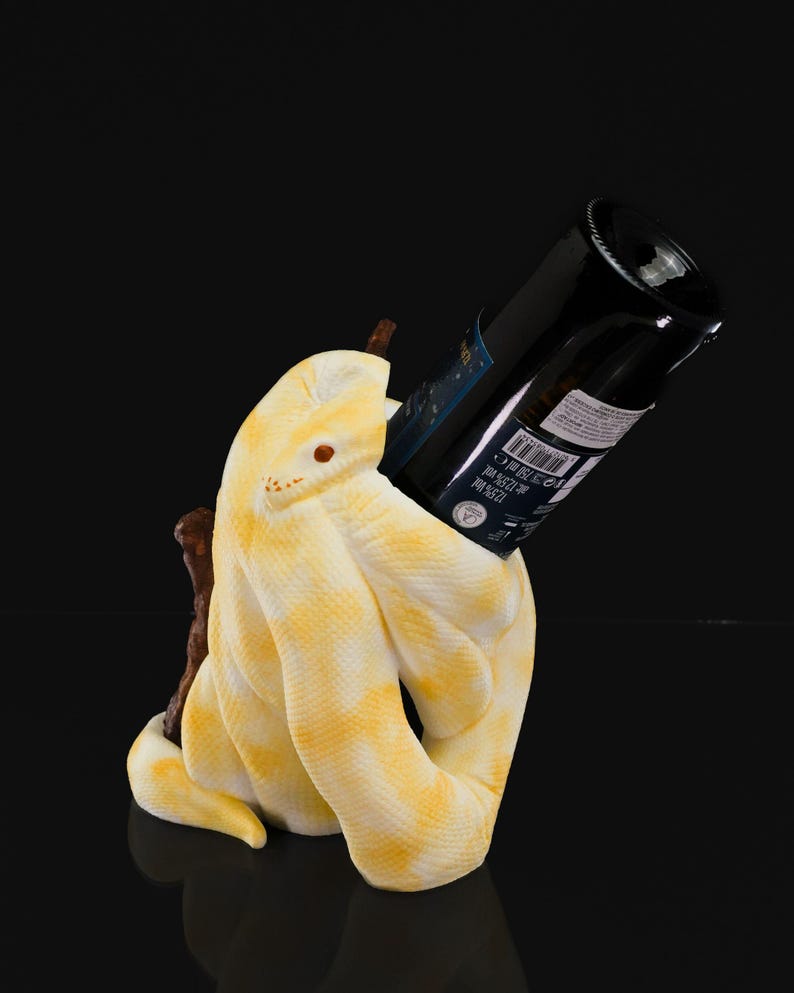 May include: A yellow and white snake-shaped wine bottle holder. The holder is designed to cradle a wine bottle, with the bottle angled upwards. The snake has a detailed scale pattern and a brown accent piece.