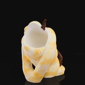 May include: A ceramic sculpture of a coiled snake in yellow and white with a brown accent. The snake's body is wrapped around itself, creating a unique and artistic form. The sculpture is set against a black background.