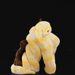 May include: A yellow and white snake sculpture with a brown branch. The snake has a textured pattern and red eyes. The sculpture is set against a black background.