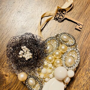 May include: A heart-shaped ornament made of white pearls, beads, and a wire flower. The ornament is decorated with a gold key and a ribbon.