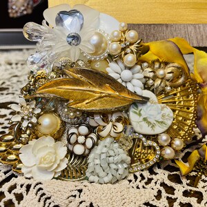 May include: A heart-shaped brooch made of gold-toned metal and white and gold-toned plastic flowers, pearls, and rhinestones. The brooch is decorated with a gold-toned leaf and a white porcelain flower with a green stem.
