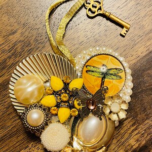 May include: A heart-shaped ornament made of gold and white beads, buttons, and other embellishments. The ornament features a dragonfly, a butterfly, and a flower. A gold key is attached to the ornament.