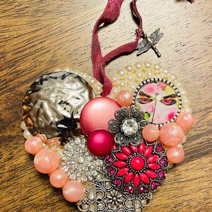 May include: A heart-shaped ornament decorated with pink, white, and silver beads, buttons, and charms. A dragonfly charm hangs from the top of the ornament.