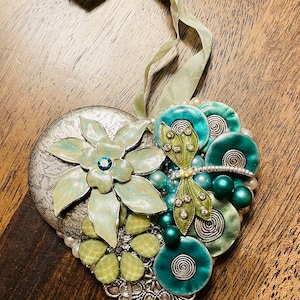 May include: A silver heart-shaped ornament decorated with green and blue flowers, beads, and a dragonfly. The ornament has a green ribbon attached to the top.