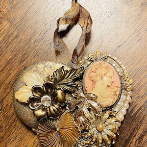 May include: A heart-shaped ornament made of gold and white beads, with a cameo of a woman's face in the center. The ornament is decorated with gold leaves and flowers.