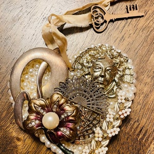 May include: A heart-shaped ornament made of white pearls and gold-toned metal accents. The ornament features a flower, a fan, a face, and a key with the number "1111" on it.