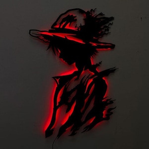 Puede incluir: Una silueta negra de un personaje con sombrero de paja, con iluminación LED roja que delinea la figura. La obra de arte está montada en una pared gris, creando un contraste llamativo entre la figura oscura y los detalles iluminados.