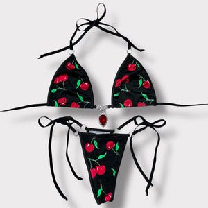May include: Black bikini set with a cherry print. The triangle top and thong-style bottom feature a pattern of red cherries and green leaves. A silver-toned charm with a red gem adorns the top. The bikini has black ties.