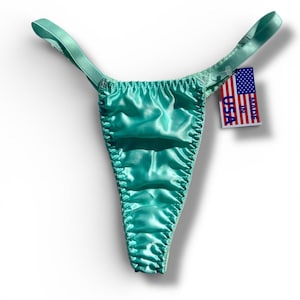 May include: A turquoise satin thong with delicate straps. The garment has a ruched front and a tag that reads "Made in USA."
