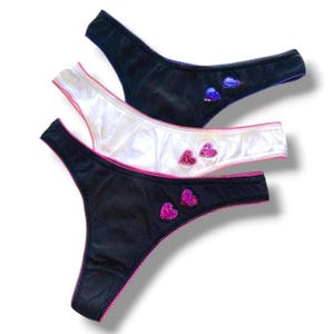 Puede incluir: Tres tangas negros y blancos. Cada tanga tiene un ribete rosa y adornos en forma de corazón. Los tangas negros tienen corazones rosas y morados, mientras que el tanga blanco tiene corazones rosas.
