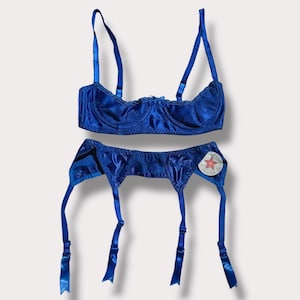 May include: A two-piece lingerie set in a vibrant blue hue. The set includes a bra with adjustable straps and a garter belt. The fabric has a subtle floral pattern. A tag with a star and the text "MADE IN USA" is attached to the garter belt.