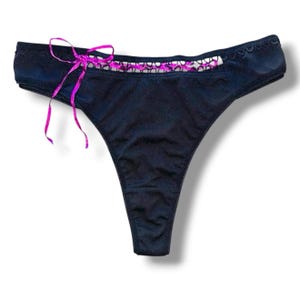 Puede incluir: Tanga negro con un lazo de cinta rosa en el lado izquierdo. El borde superior presenta un ribete festoneado y detalles de encaje rosa y blanco. El tanga está hecho de una tela suave y opaca.