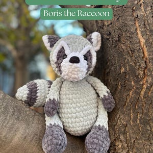 May include: A crocheted plush raccoon toy, in shades of gray, white, and black, perched on a tree branch. The raccoon has a striped tail and a distinctive mask around its eyes. The text "Pattern PDF Only" and "Boris the Raccoon" are visible.